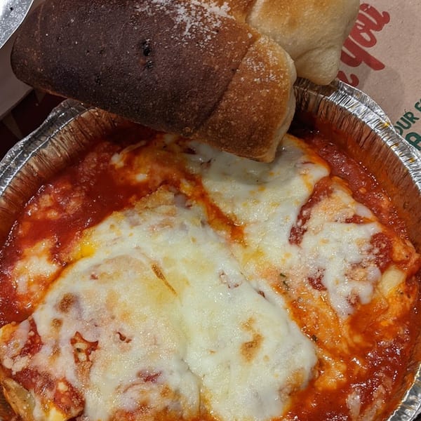 Baked pasta with melted cheese and red sauce in a foil container, with breadsticks on top.